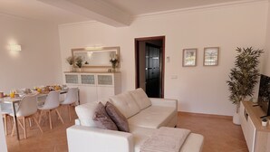 Ein Wohnzimmer mit einem weißen Sofa, einem Essbereich mit Stühlen, einer Küchenzeile mit Schränken und einer großen Pflanze.