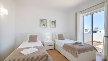 A hotel room with two single beds, a bedside table with a lamp, and a large sliding glass door leading to a balcony.