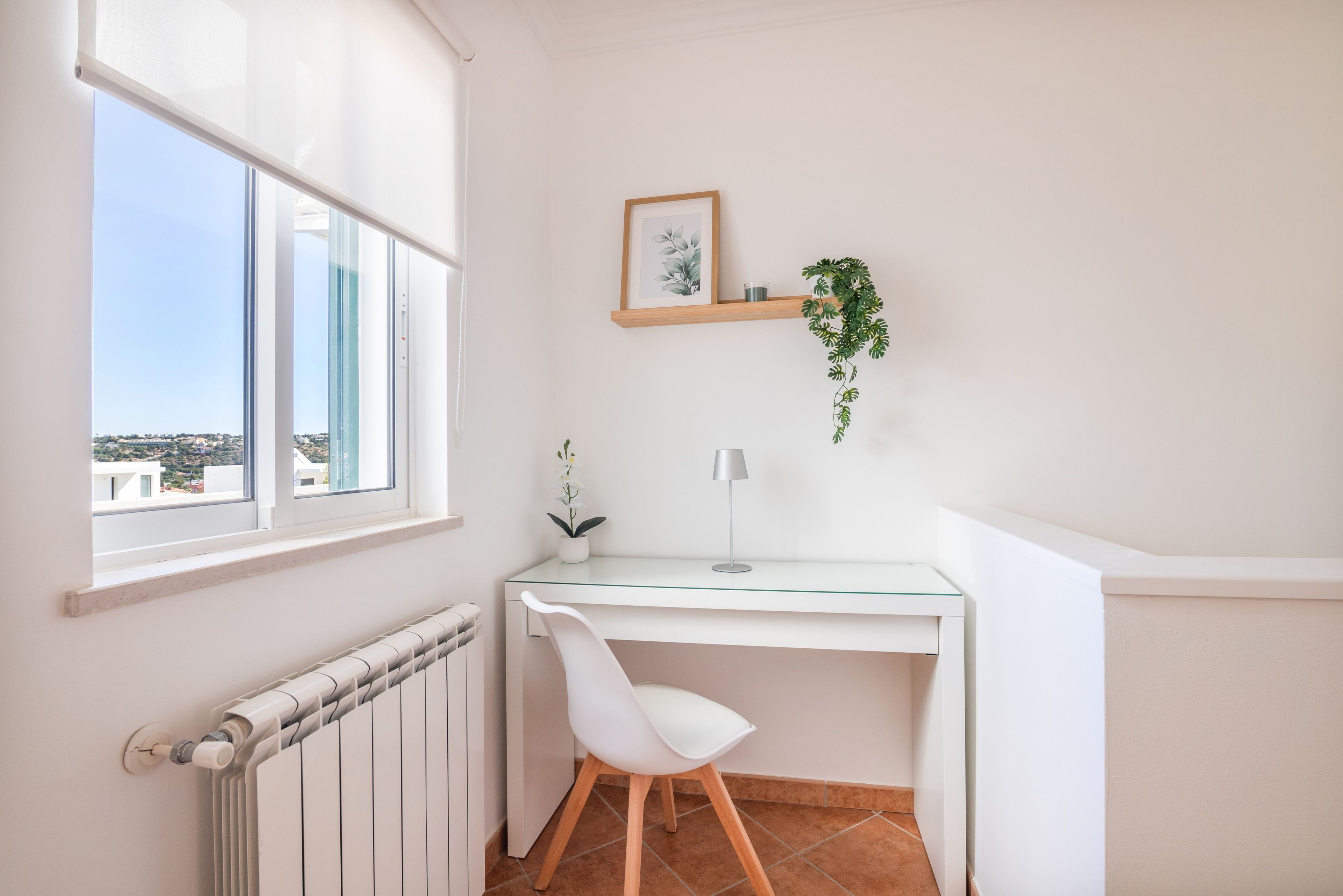 Een hoekappartement met een wit bureau, een stoel, een radiator, een raam met uitzicht, een ingelijst kunstwerk en een hangende plant.