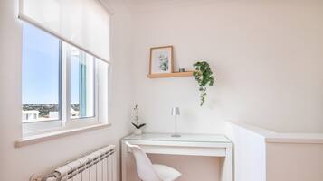 Een hoekappartement met een wit bureau, een stoel, een radiator, een raam met uitzicht, een ingelijst kunstwerk en een hangende plant.