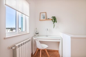 Ett hörnrum med ett vitt skrivbord, en stol, ett element, ett fönster med utsikt, en inramad tavla och en hängande växt.