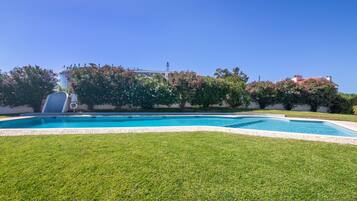 Una piscina con cielo azul despejado, rodeada de césped cuidado y arbustos florecientes.