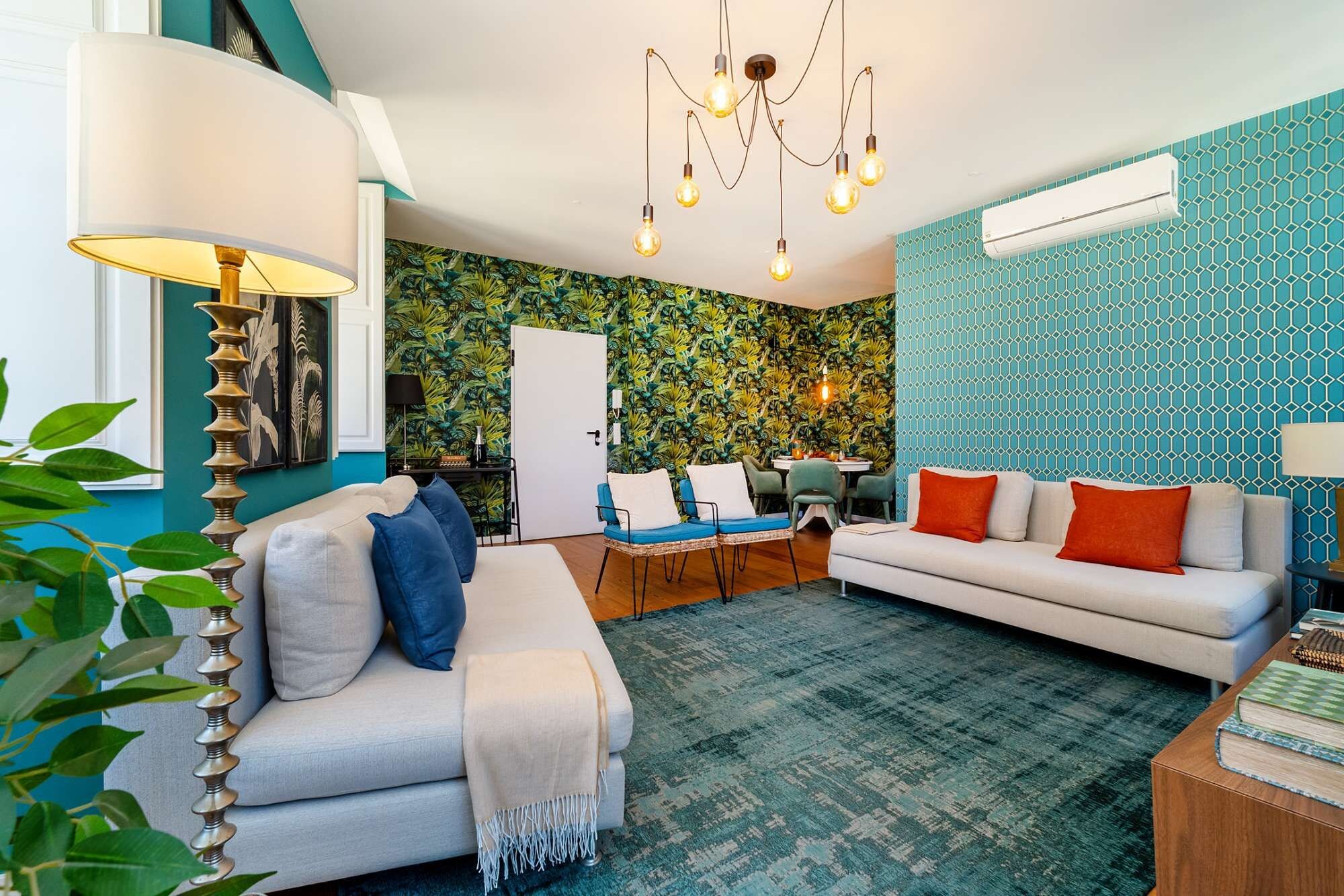 A living room with a sofa, a dining table, and chairs. There is a chandelier, a lamp, and a wall air conditioner.