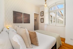 Ein Schlafzimmer mit einem Bett, Kissen, einem Nachttisch mit Lampe, gerahmten Bildern an der Wand und einem Fenster mit Blick auf Gebäude.