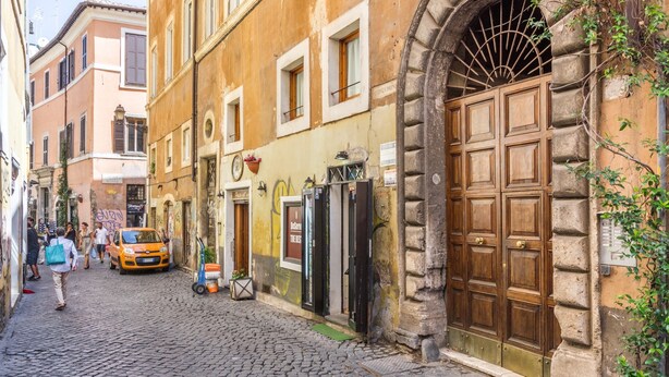 A cobblestone street with historic buildings, a large wooden door, and a parked car. - Secret Loft in Trastevere - Moro (Rome)