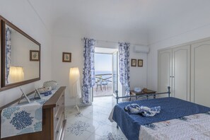 Ein Schlafzimmer mit Bett, Kommode, Schreibtisch und Stuhl. Ein Fenster mit Vorhängen bietet Blick auf das Meer.
