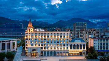 A hotel with a prominent tower and arched windows, surrounded by modern buildings and mountains in the background.