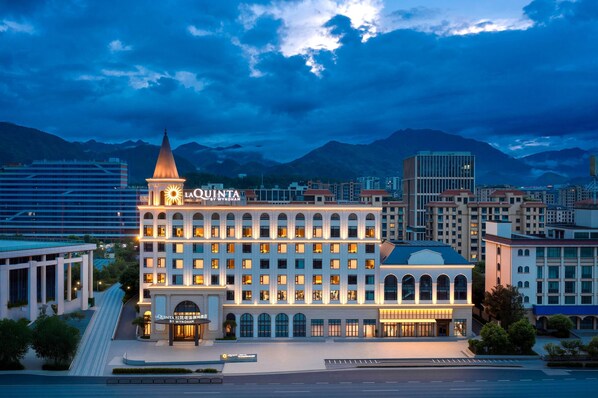 A hotel with a prominent tower and arched windows, surrounded by modern buildings and mountains in the background.