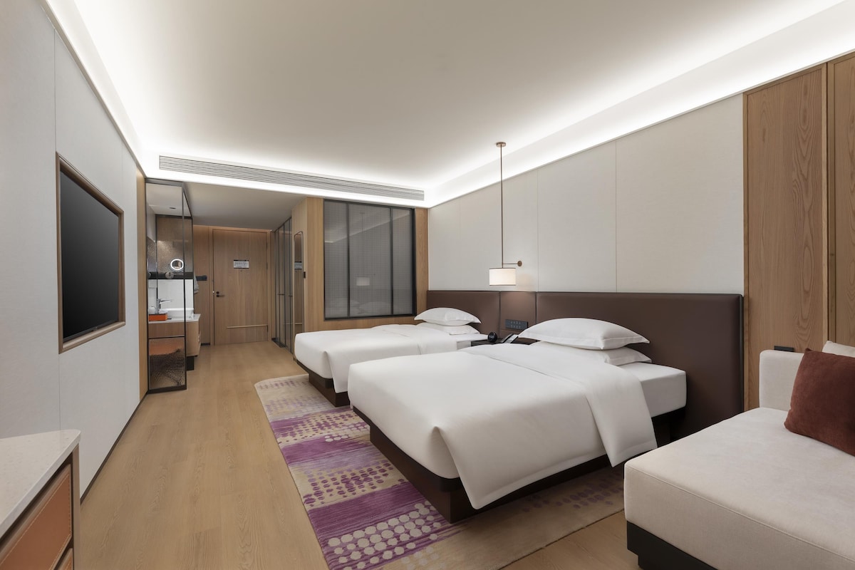 Quarto de hotel com duas camas, uma escrivaninha, um espelho, banheiro e uma janela grande.