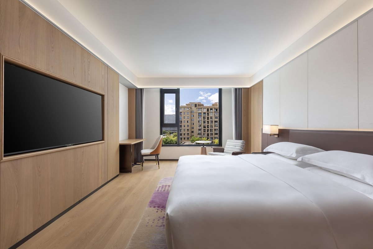 Quarto de hotel com uma cama grande, uma escrivaninha, uma cadeira, uma televisão e uma janela com vista para prédios.