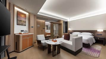 A modern hotel room with a sofa, a small table, a TV, a desk with a chair, a bed, and a bathroom visible through a glass door.