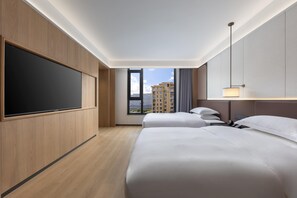 Quarto de hotel com duas camas, televisão de tela plana, janela vista para a cidade e luminária moderna.