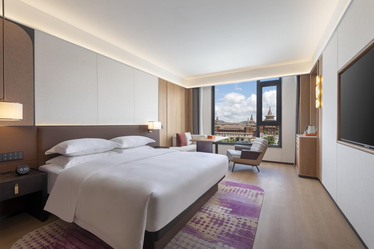 Quarto de hotel com cama, mesa de cabeceira, abajur e vista para prédios por uma janela grande.