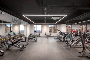Un gimnasio moderno con varios equipos de ejercicio, ventanas amplias y vistas a edificios.