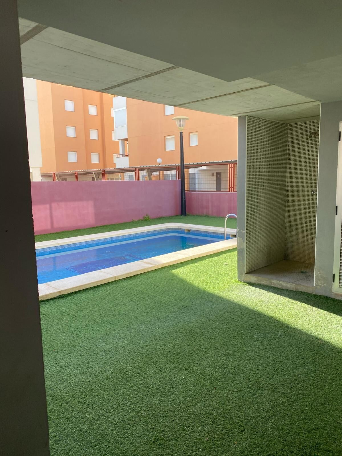 A swimming pool with artificial grass, a covered area, and a building with orange walls in the background.