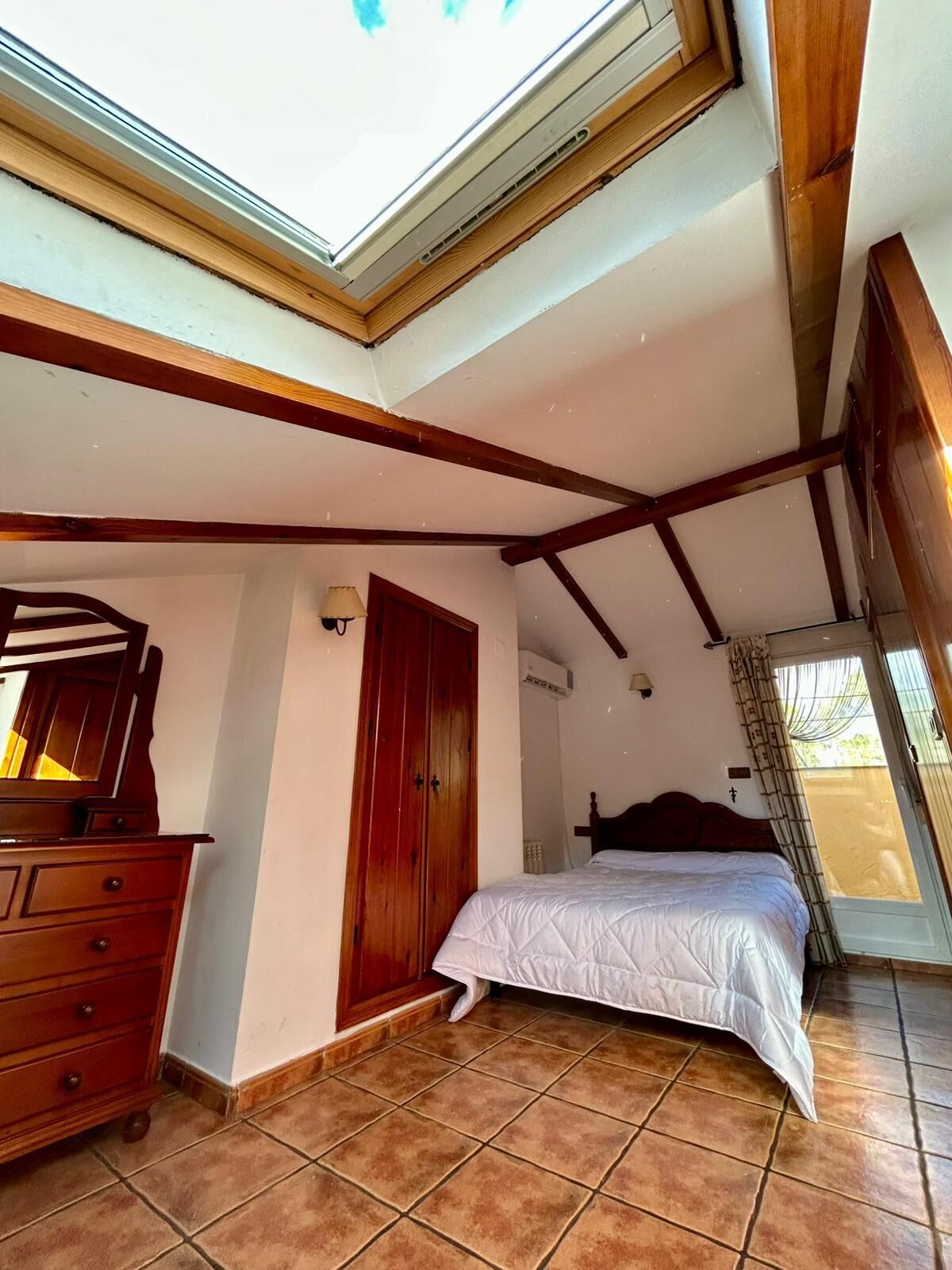Un dormitorio con tragaluz, vigas de madera, una cama, un aparador y una puerta.