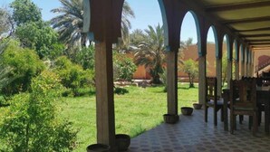 An outdoor patio area with a covered seating area, surrounded by lush greenery and palm trees. - Gite DAR UL (Er-Rteb)