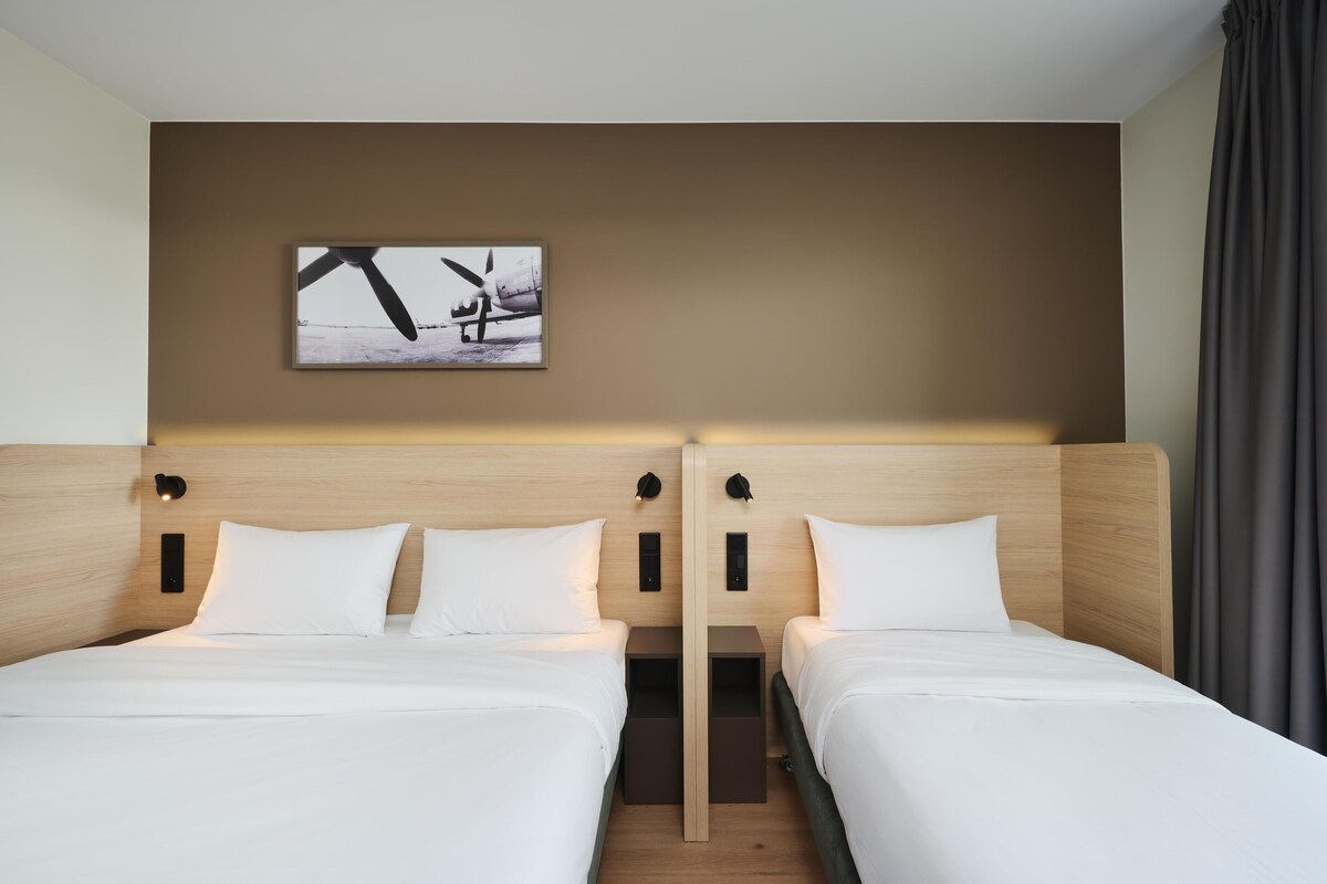 A hotel room with two beds, a headboard with integrated lighting, a wall-mounted artwork, and a curtain.