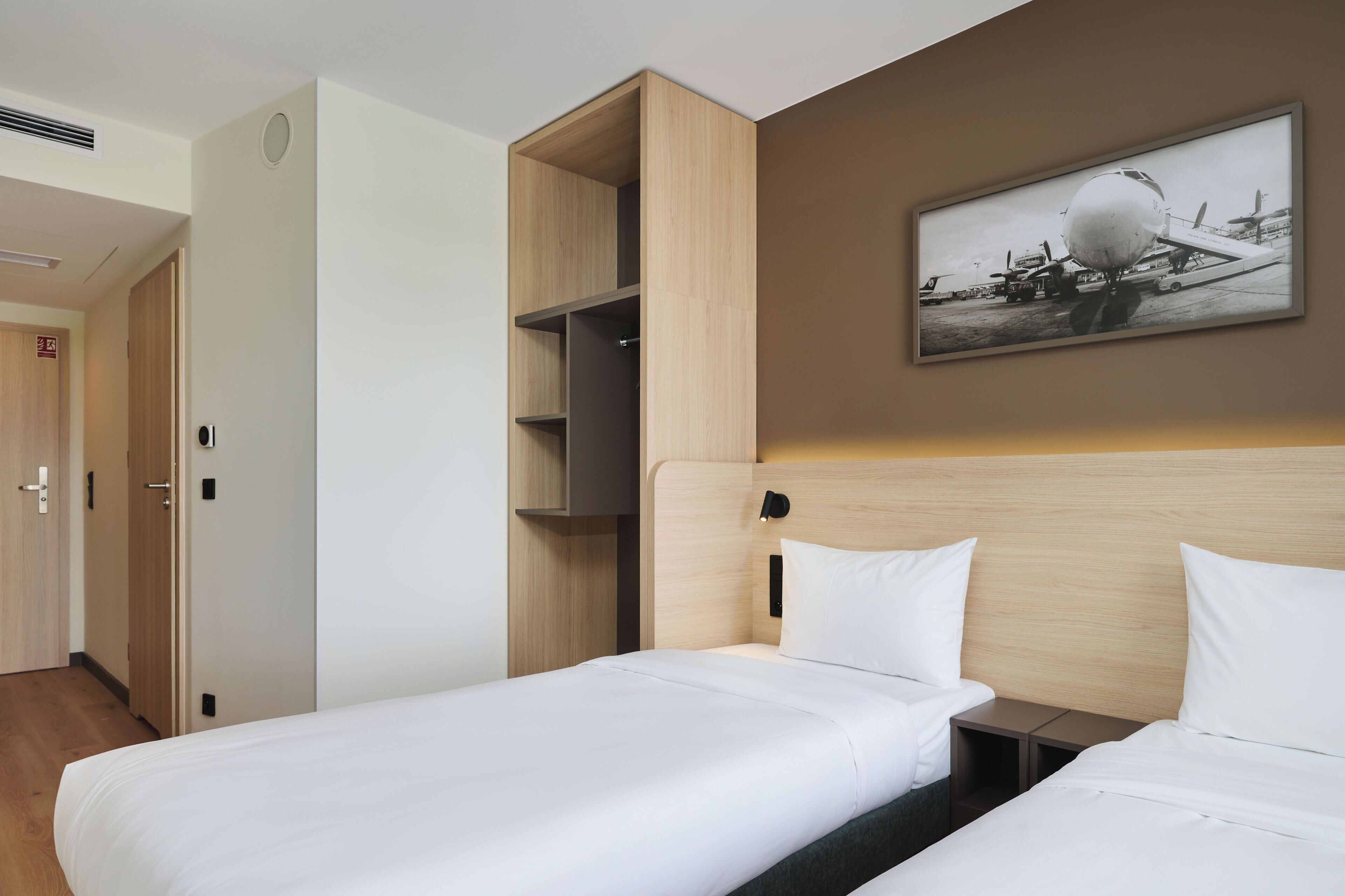 A hotel room with a bed, a nightstand, a wooden wardrobe, a wall-mounted shelf, and a framed picture of an airplane.