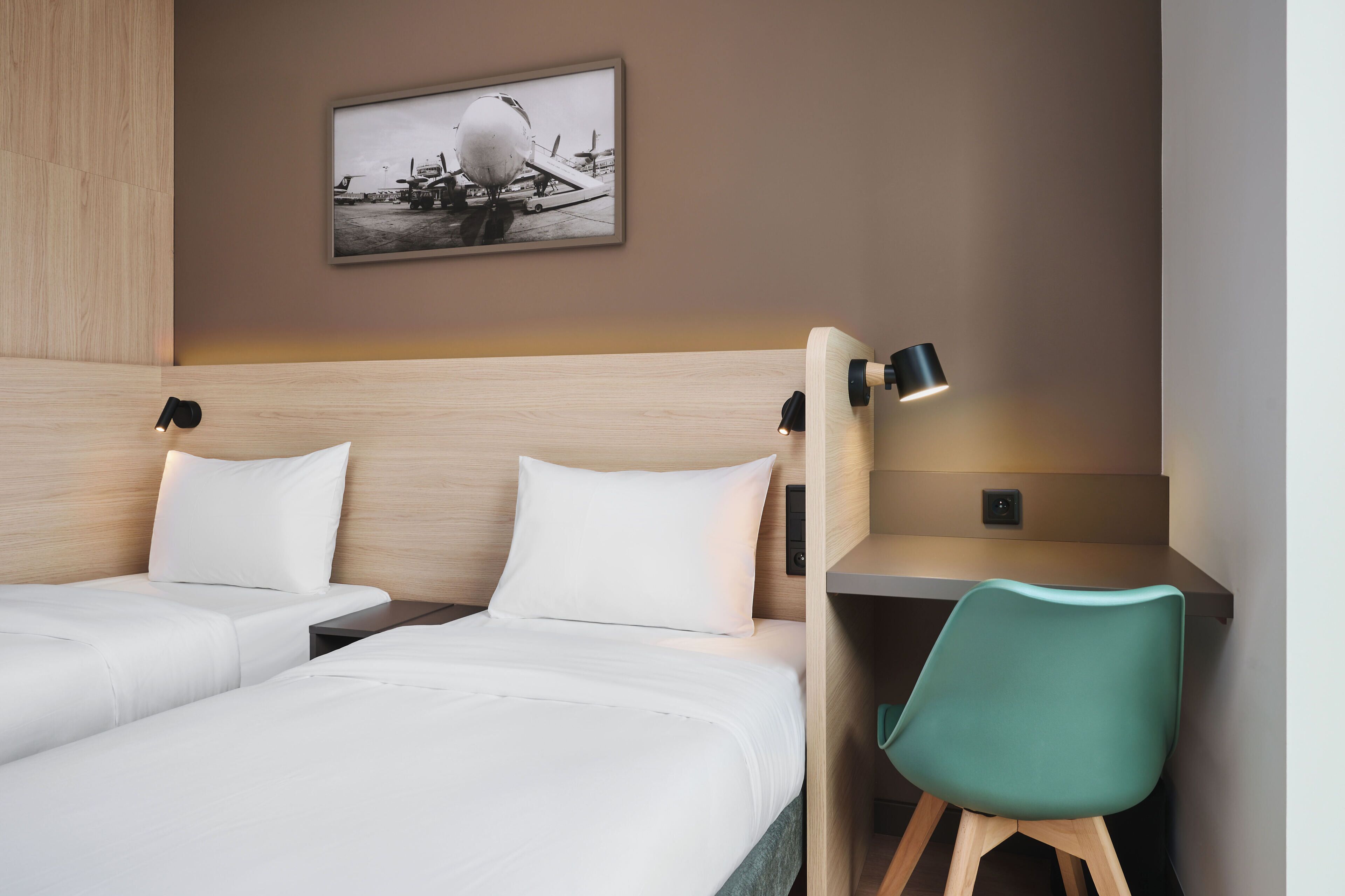 A hotel room with two beds, a desk, a chair, a wall-mounted picture, and a nightstand with a lamp.