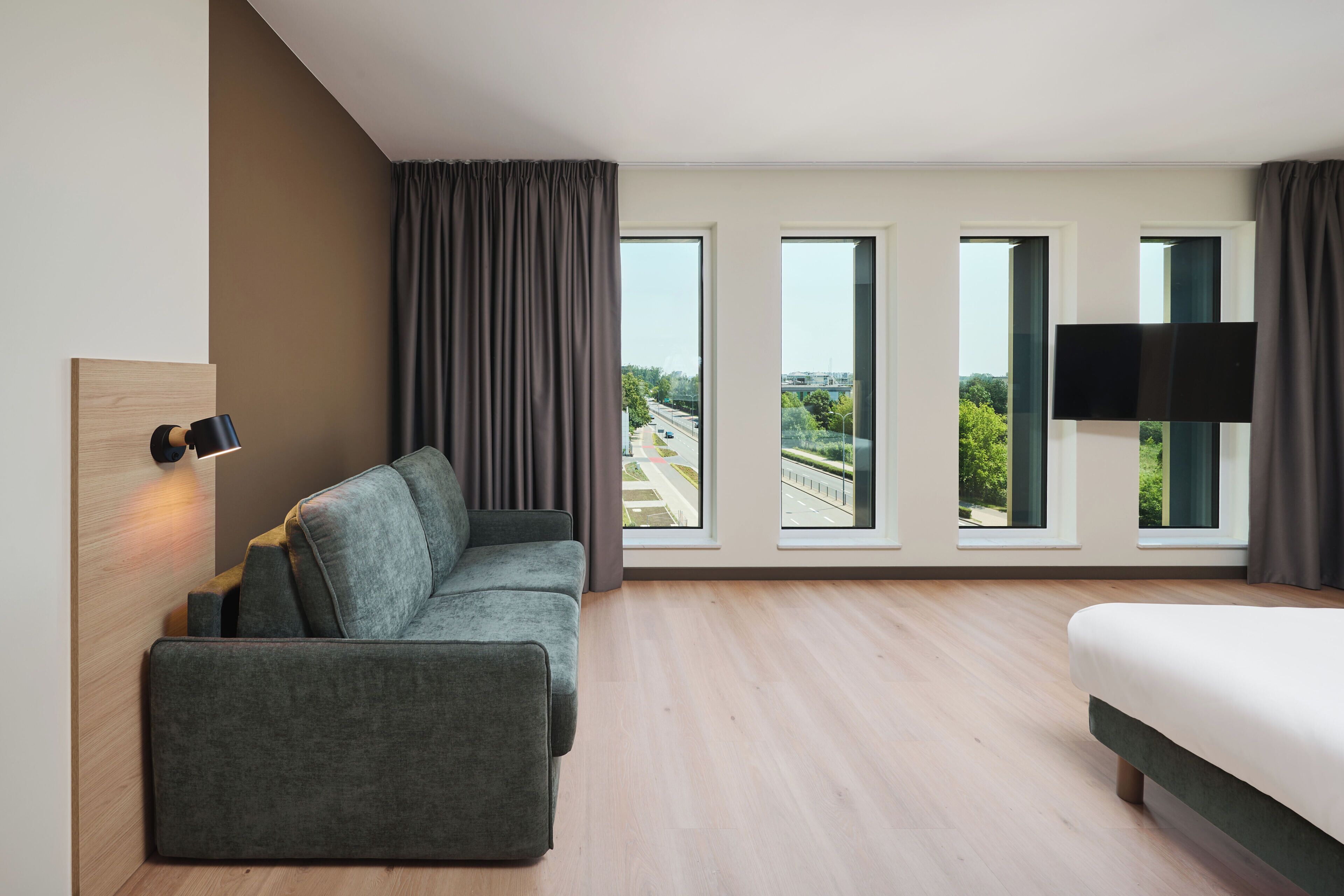 A modern hotel room with a sofa, a bed, a flat-screen TV, and large windows offering a city view.