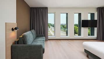 A modern hotel room with a sofa, a bed, a flat-screen TV, and large windows offering a city view.