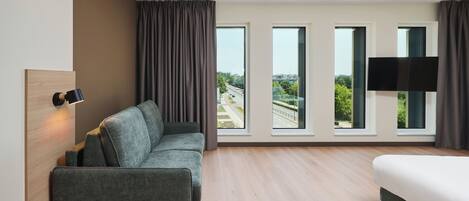 Une chambre d’hôtel moderne dotée d’un canapé, d’un lit, d’un téléviseur à écran plat et de grandes fenêtres offrant une vue sur la ville.