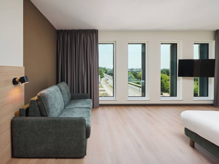 A modern hotel room with a sofa, a bed, a flat-screen TV, and large windows offering a city view.