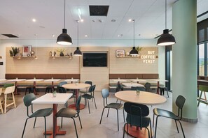 Un interior de cafetería moderna con mesas redondas, sillas, iluminación colgante y un televisor montado en la pared.