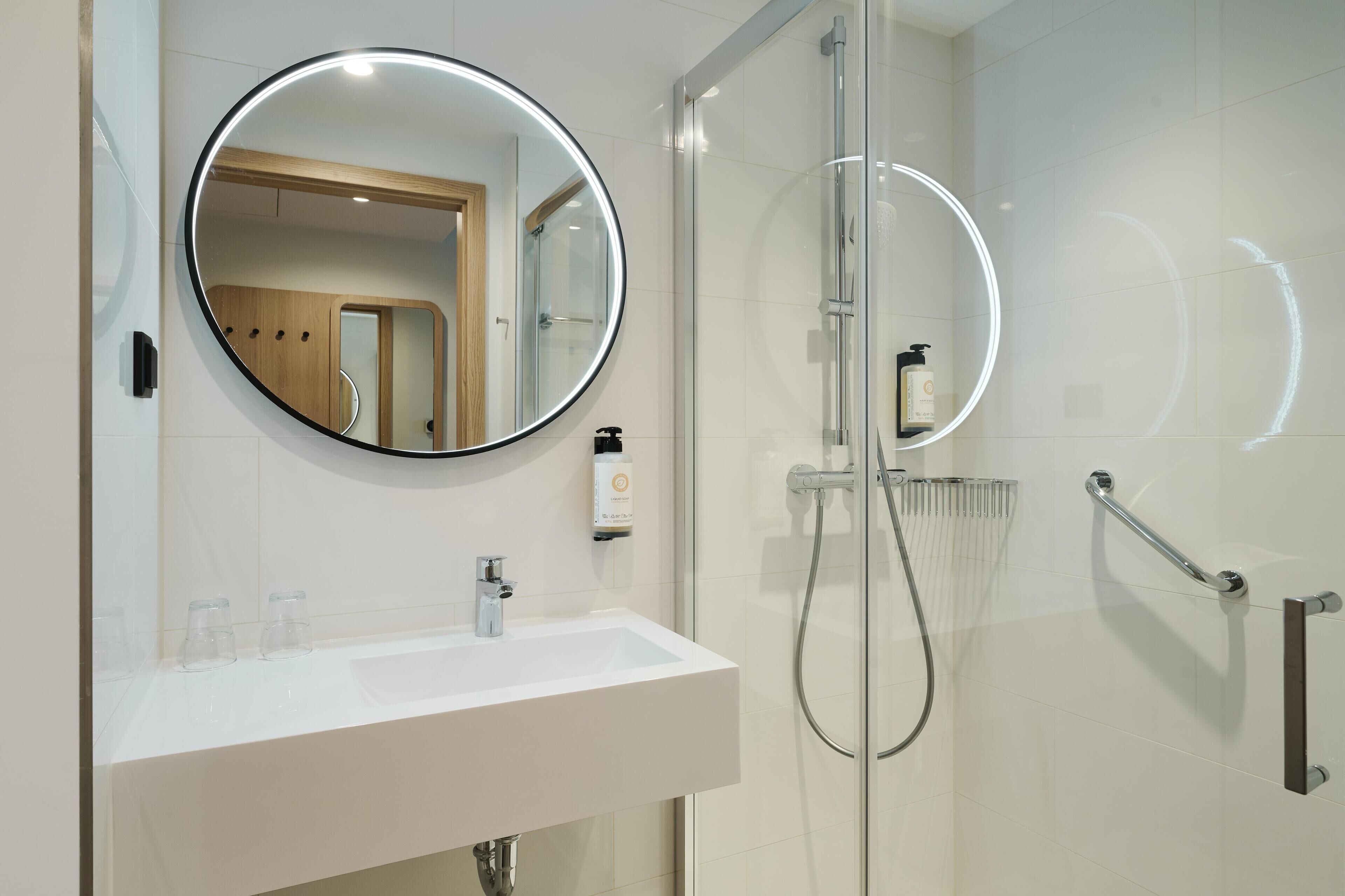 A modern bathroom with a glass-enclosed shower, a round mirror, and a white sink.