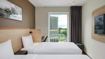Une chambre d’hôtel avec deux lits, un bureau, une chaise, une grande fenêtre donnant sur des immeubles et un téléviseur fixé au mur.