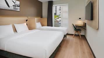 Ein Hotelzimmer mit zwei Betten, einem Schreibtisch, einem Sessel, einem Fernseher und einem Fenster mit Blick auf Gebäude.