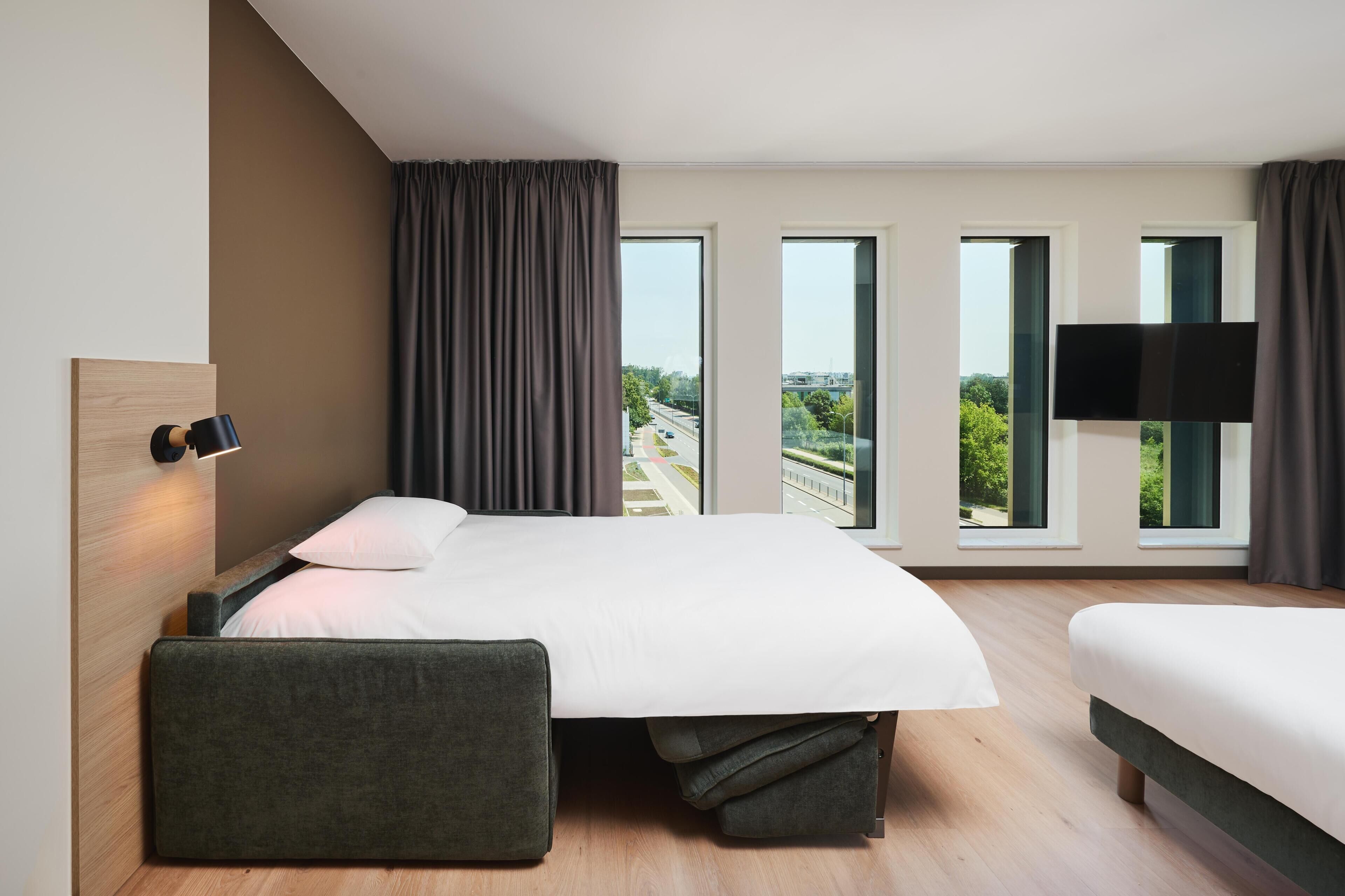 Habitación de hotel moderna con una cama grande, una mesita de noche, un televisor de pantalla plana y vistas a la ciudad a través de las ventanas.