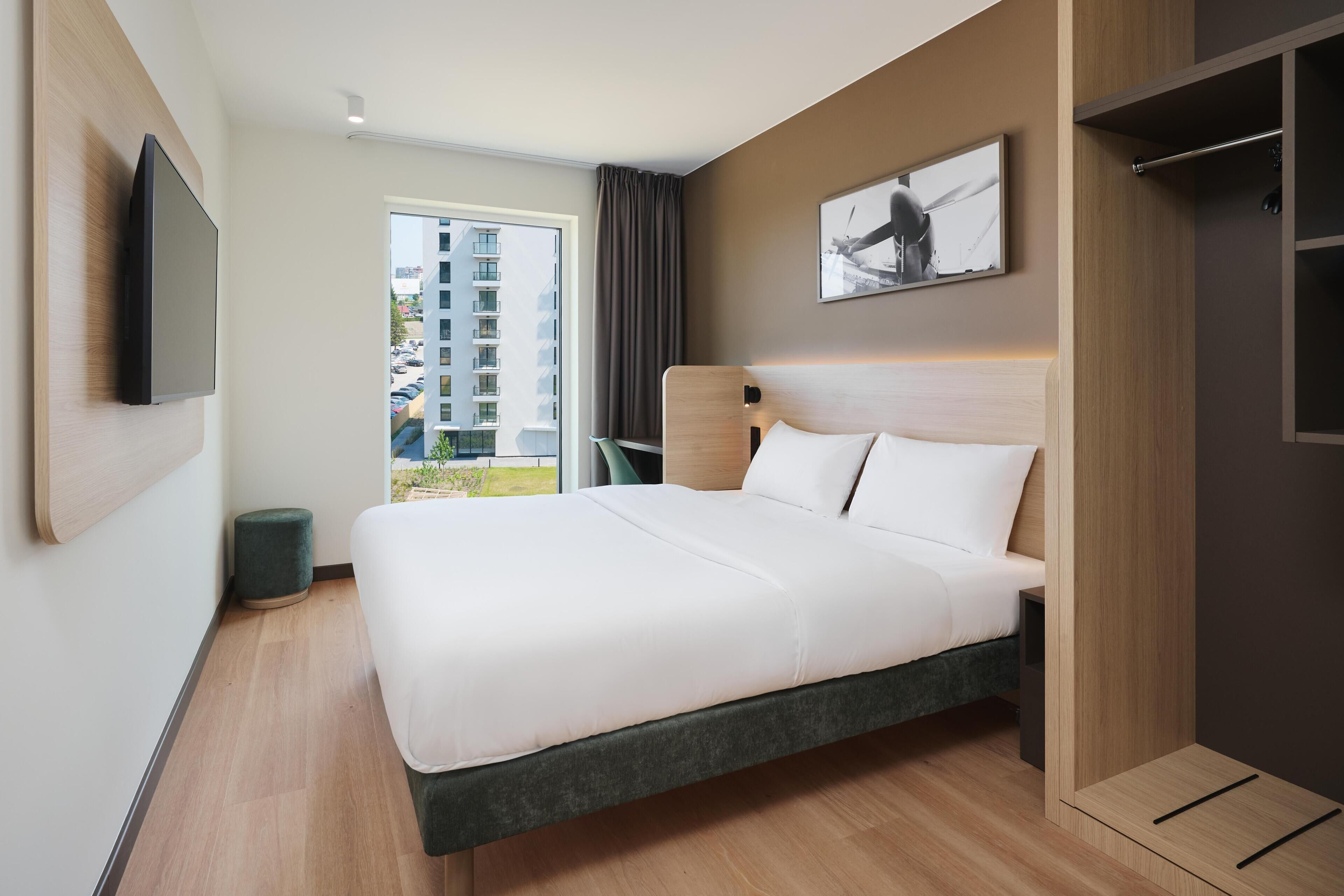 A modern hotel room with a large bed, a flat-screen TV mounted on the wall, a window with a view of a building, and a small round stool.