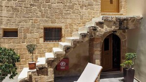 Un edificio de piedra con una escalera que lleva a una puerta, una silla de descanso, una mesa con comida y una bebida, y un jarrón con flores.
