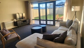 A living room with a sofa, armchair, coffee table, TV, and a view through large windows. - Urban Retreat in the CBD (Blenheim)