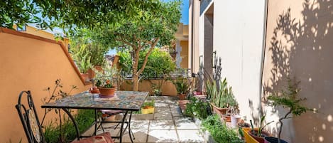 A narrow outdoor patio with a table and chairs, surrounded by potted plants and a tree.