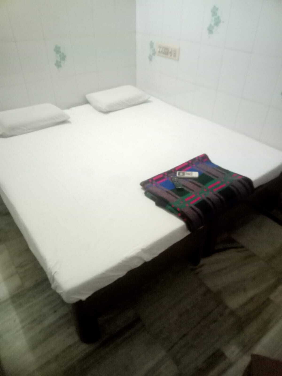 A single bed with a white mattress and pillows, a blanket, and a small bedside table with a lamp.