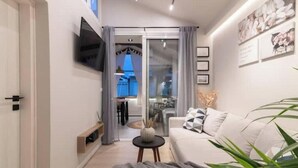 A modern living room with a sofa, a small side table, a TV mounted on the wall, and a glass door leading to a dining area. - Chrison modern house (Zakynthos)
