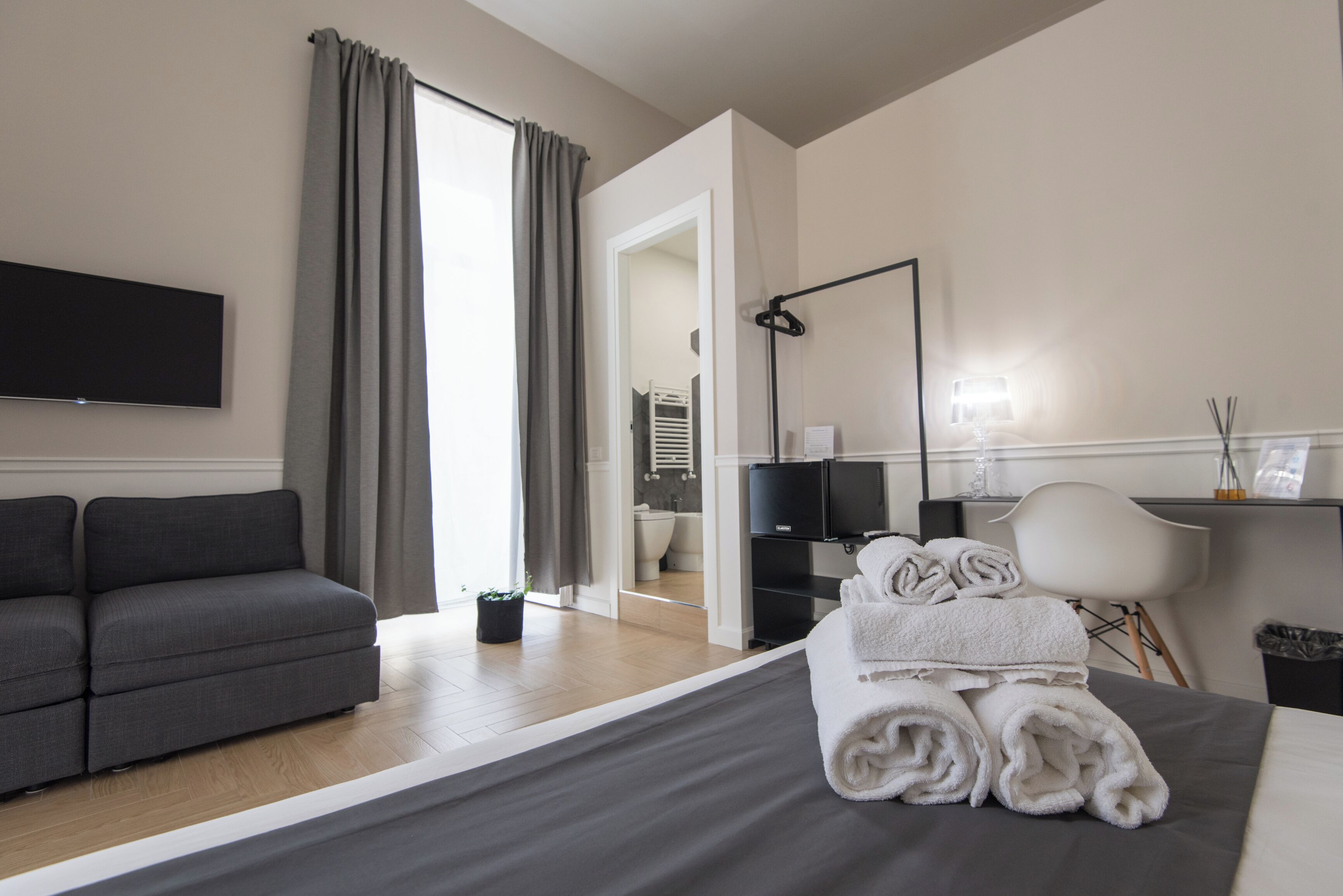A modern hotel room with a grey sofa, a neatly made bed with rolled towels, a flat-screen TV, a desk with a lamp, and a view of the bathroom.