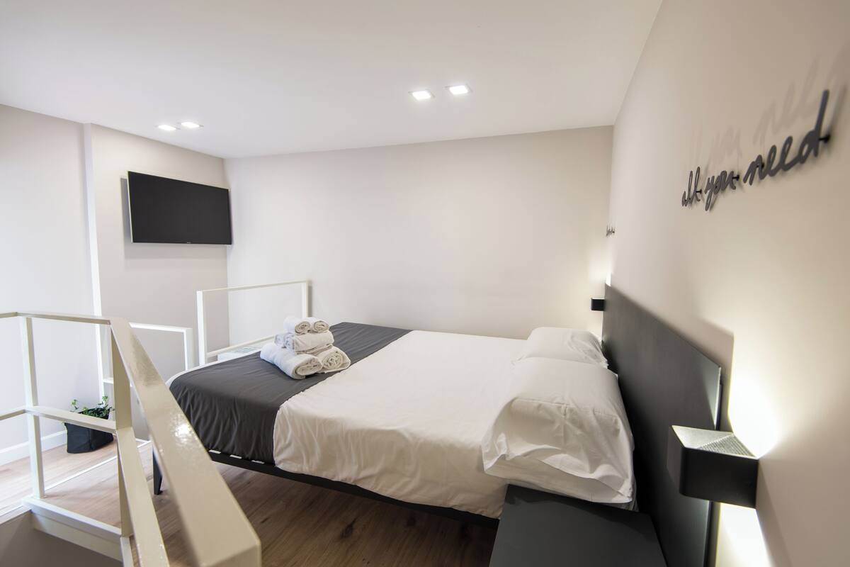 A modern hotel room with a bed, a flat-screen TV mounted on the wall, and a bedside table with a lamp.