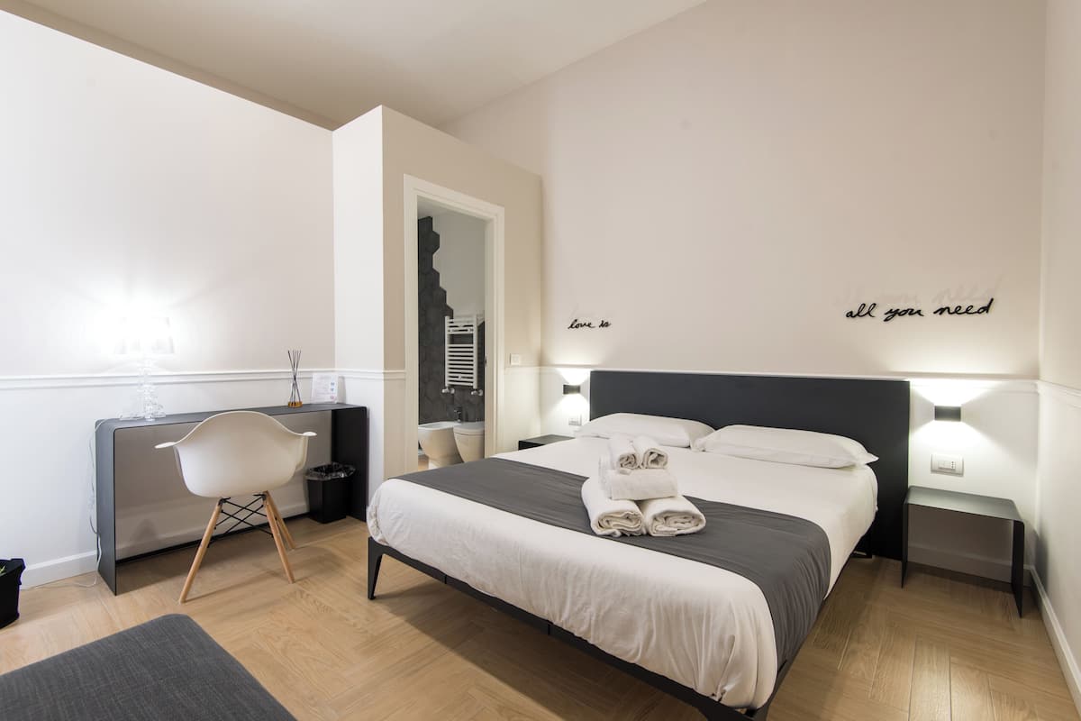 A modern hotel room with a neatly made bed, a desk with a chair, and a small bedside table.