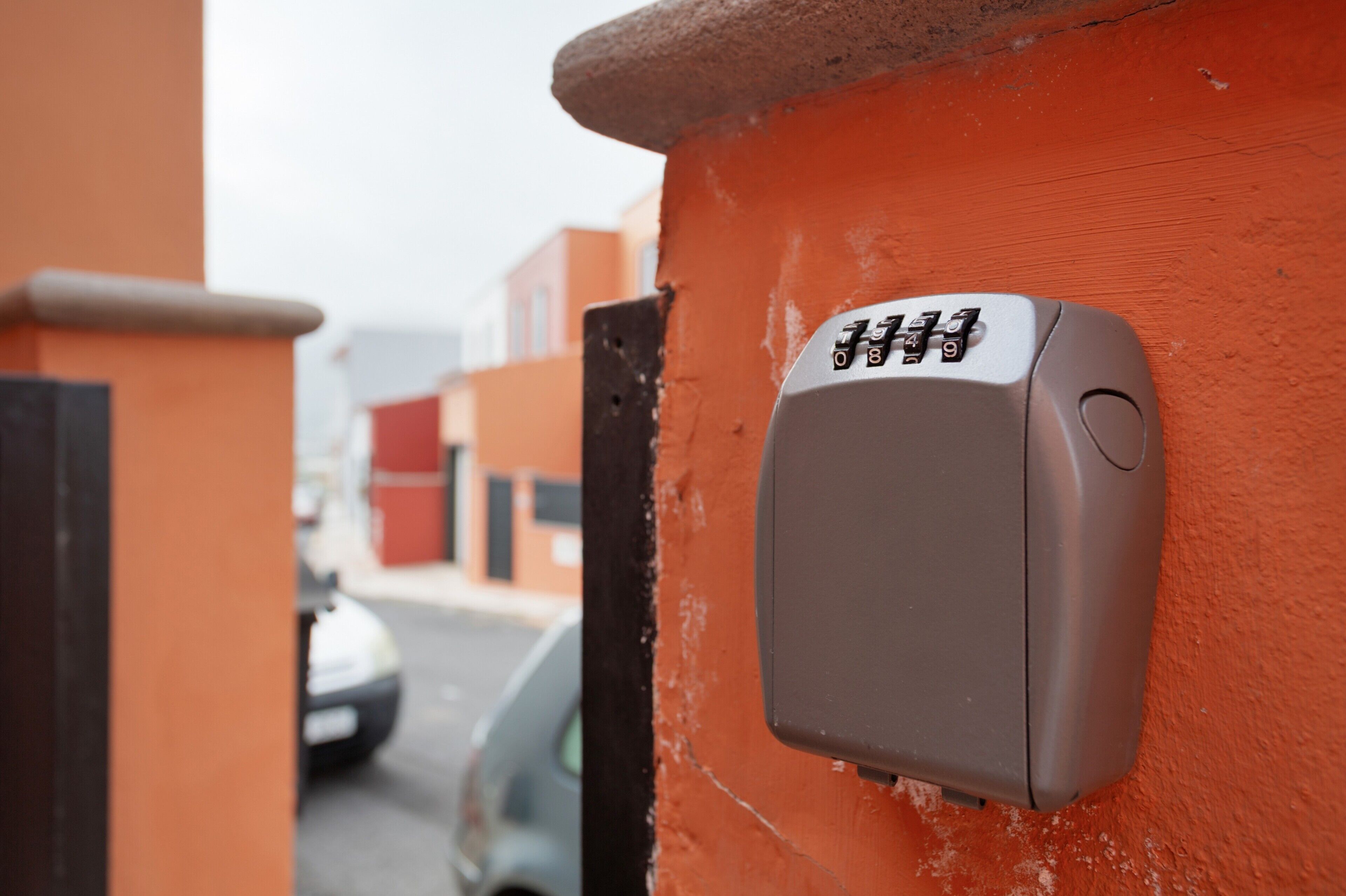 Un panel de control de seguridad adherido a una pared naranja, un automóvil estacionado al fondo y un edificio con puerta roja.