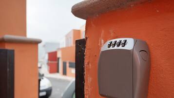 Un panel de control de seguridad adherido a una pared naranja, un automóvil estacionado al fondo y un edificio con puerta roja.