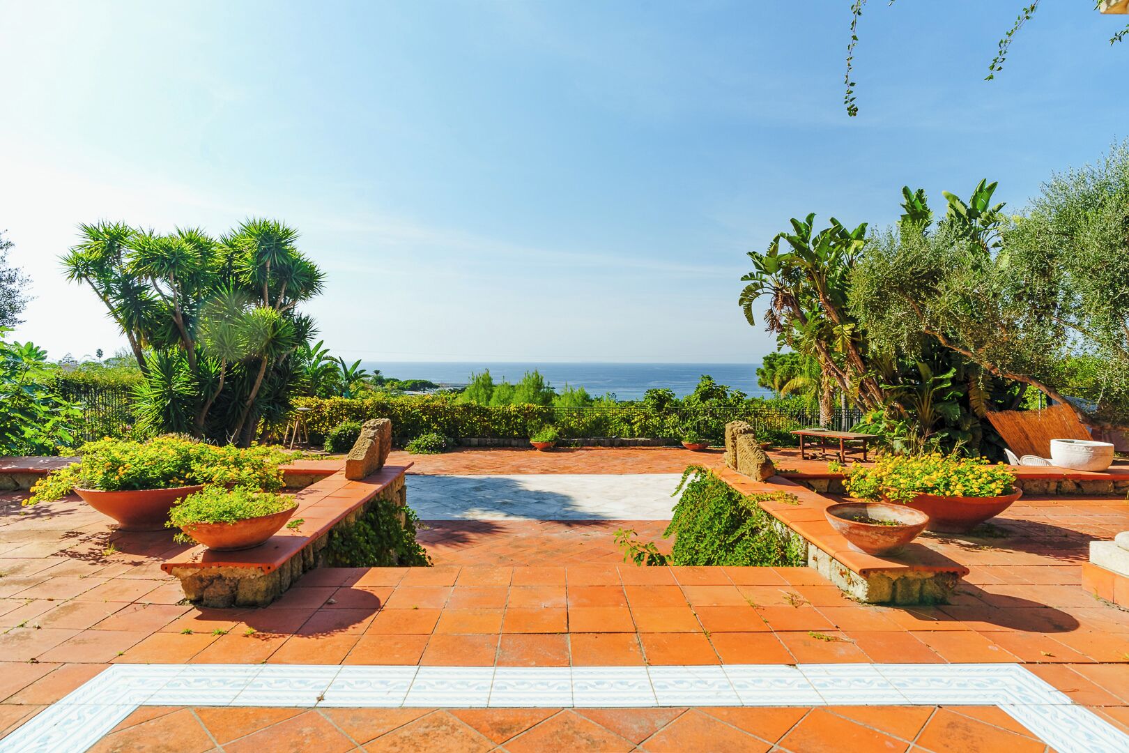 An outdoor patio with terracotta tiles, potted plants, and seating areas overlooking a body of water.