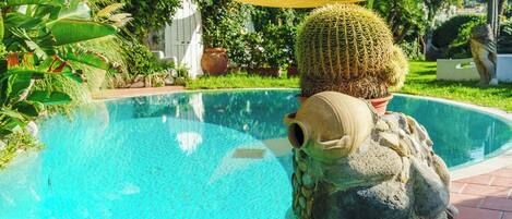 A pool with a cactus sculpture, surrounded by greenery and a tiled deck.