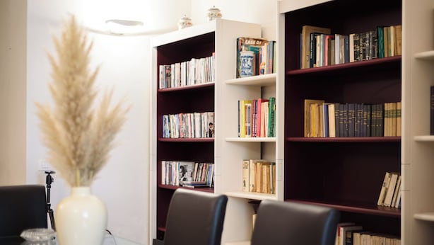 Et bibliotek med bogreoler, et bord med stole og en vase med tørrede planter.