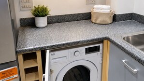 A compact kitchen with a built-in washing machine, a potted plant, and a towel holder. - Modern 2BR House Sleeps 6 Parking, Wi-Fi & Self Check-In Near Bluewater & London (Greenhithe)