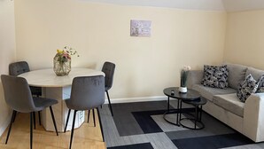 A modern living room with a round dining table, grey upholstered chairs, a grey sofa with patterned cushions, a small side table, and a black and grey rug. - Modern 2BR House Sleeps 6 Parking, Wi-Fi & Self Check-In Near Bluewater & London (Greenhithe)