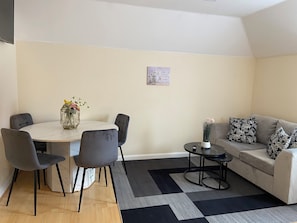 A modern living room with a round dining table, grey upholstered chairs, a grey sofa with patterned cushions, a small side table, and a black and grey rug.